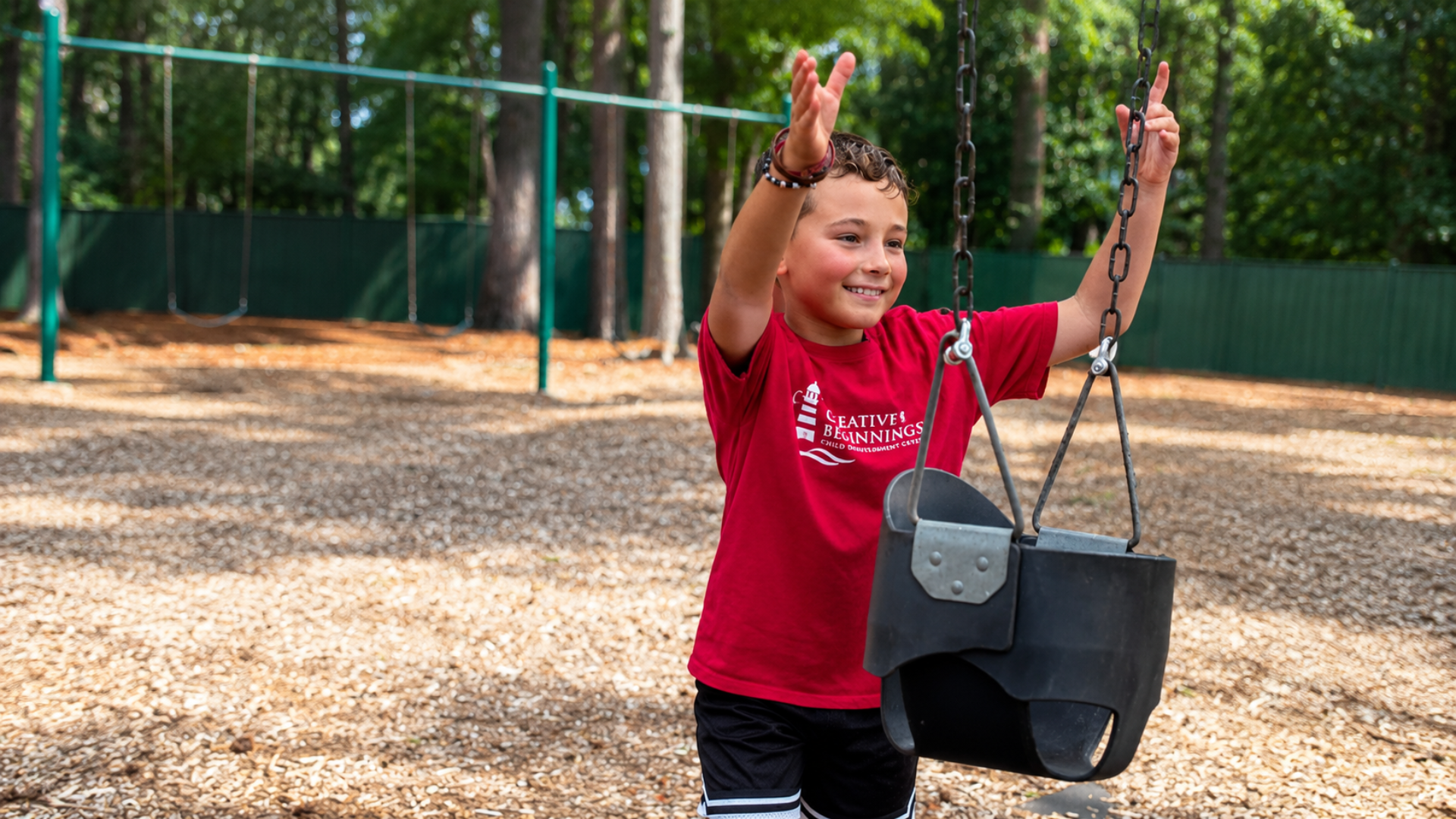 A camper in a red Creative Beginnings T-shirt cheering with both arms raised beside a swing on the shaded woodchip playground