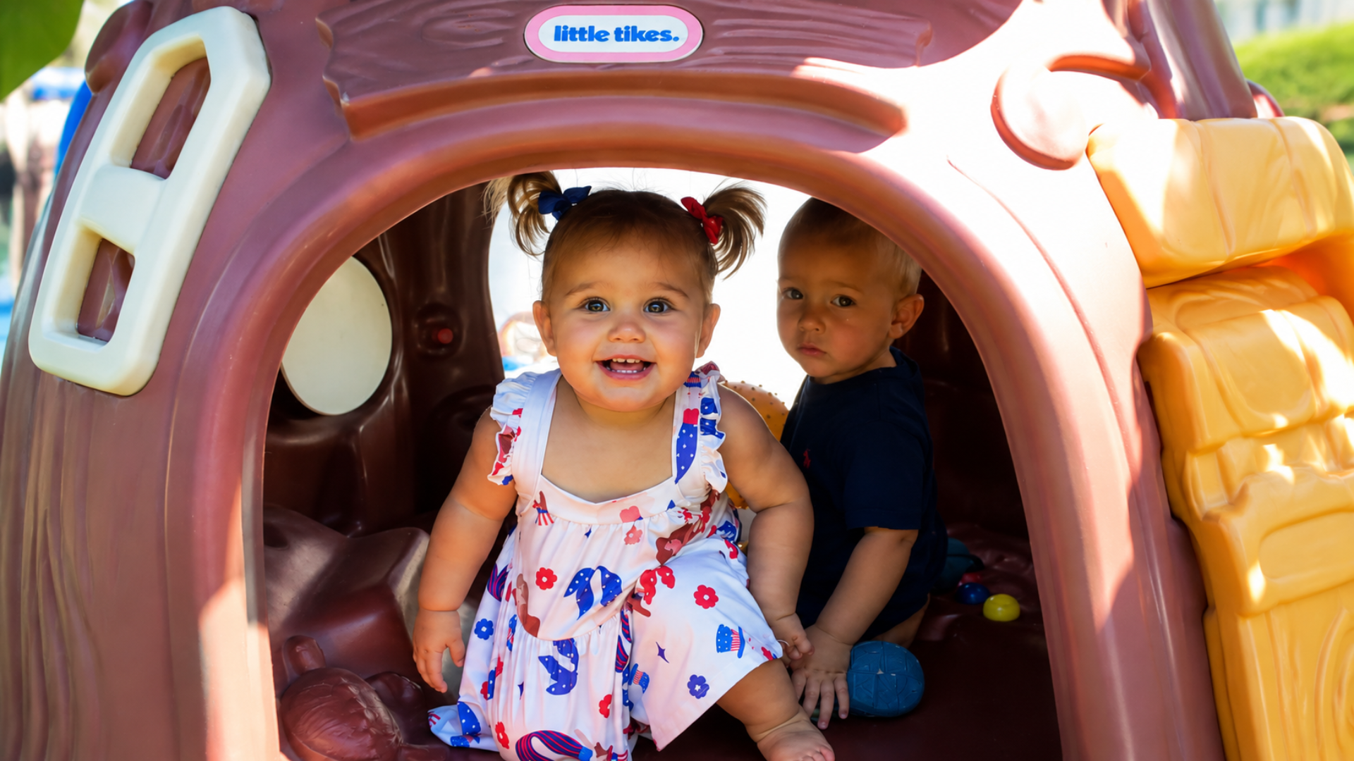Two toddlers peeking out of a plastic playhouse with a yellow slide on a sunny playground
