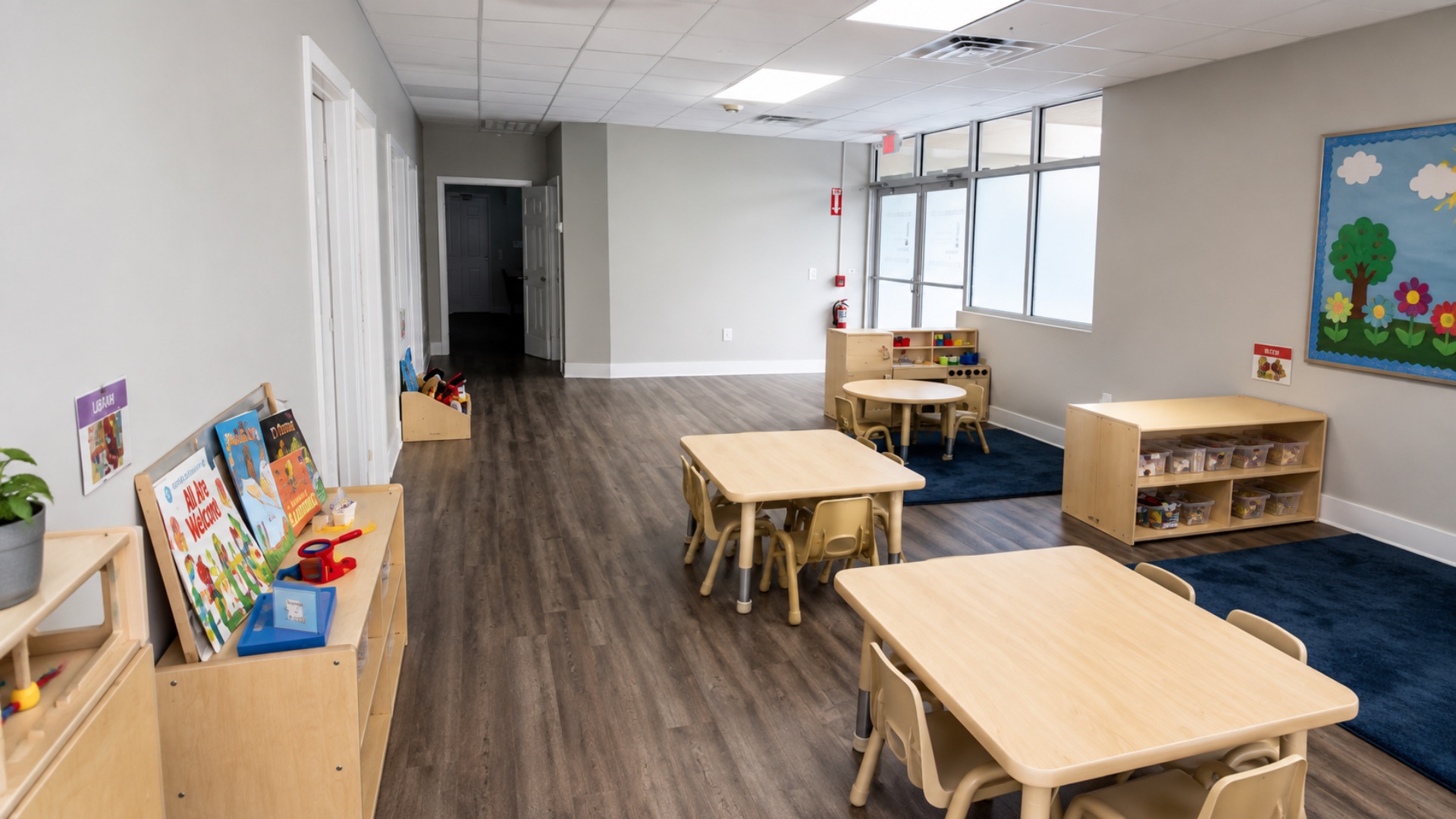 Preschool training classroom with child-height tables and chairs, a navy rug, book display and a flower and tree bulletin board on the wall