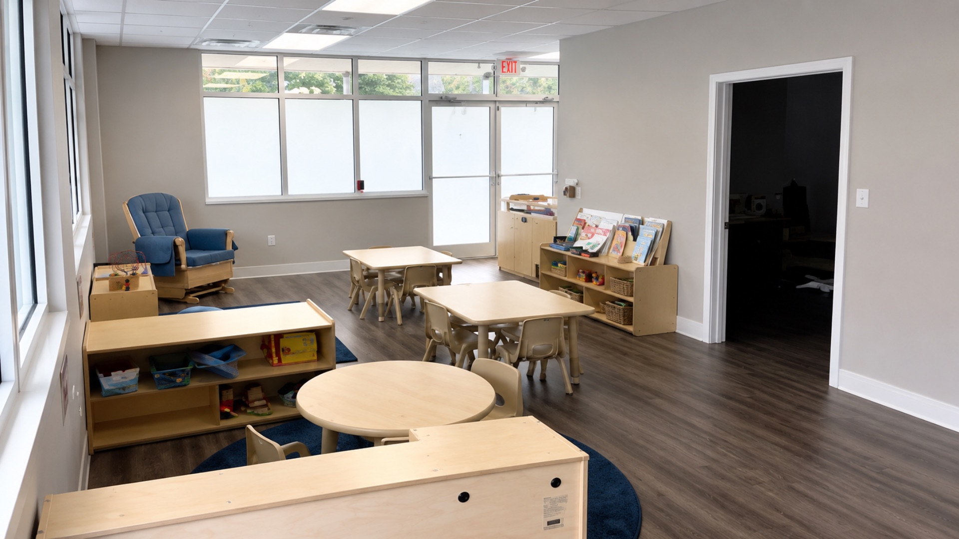 Toddler training classroom with wooden tables and chairs, a blue glider rocking chair by the windows and a library shelf of picture books