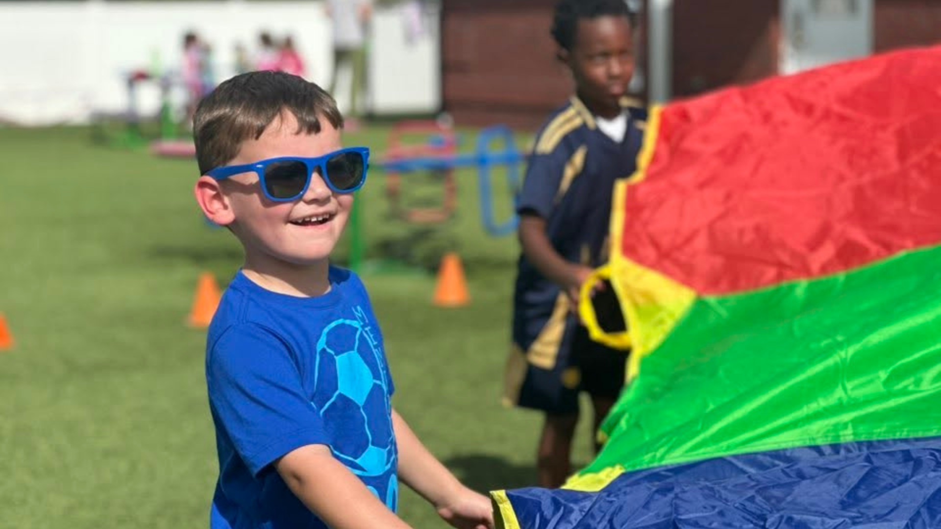 A boy in blue sunglasses and a soccer tee holding a rainbow parachute with a friend on the camp field