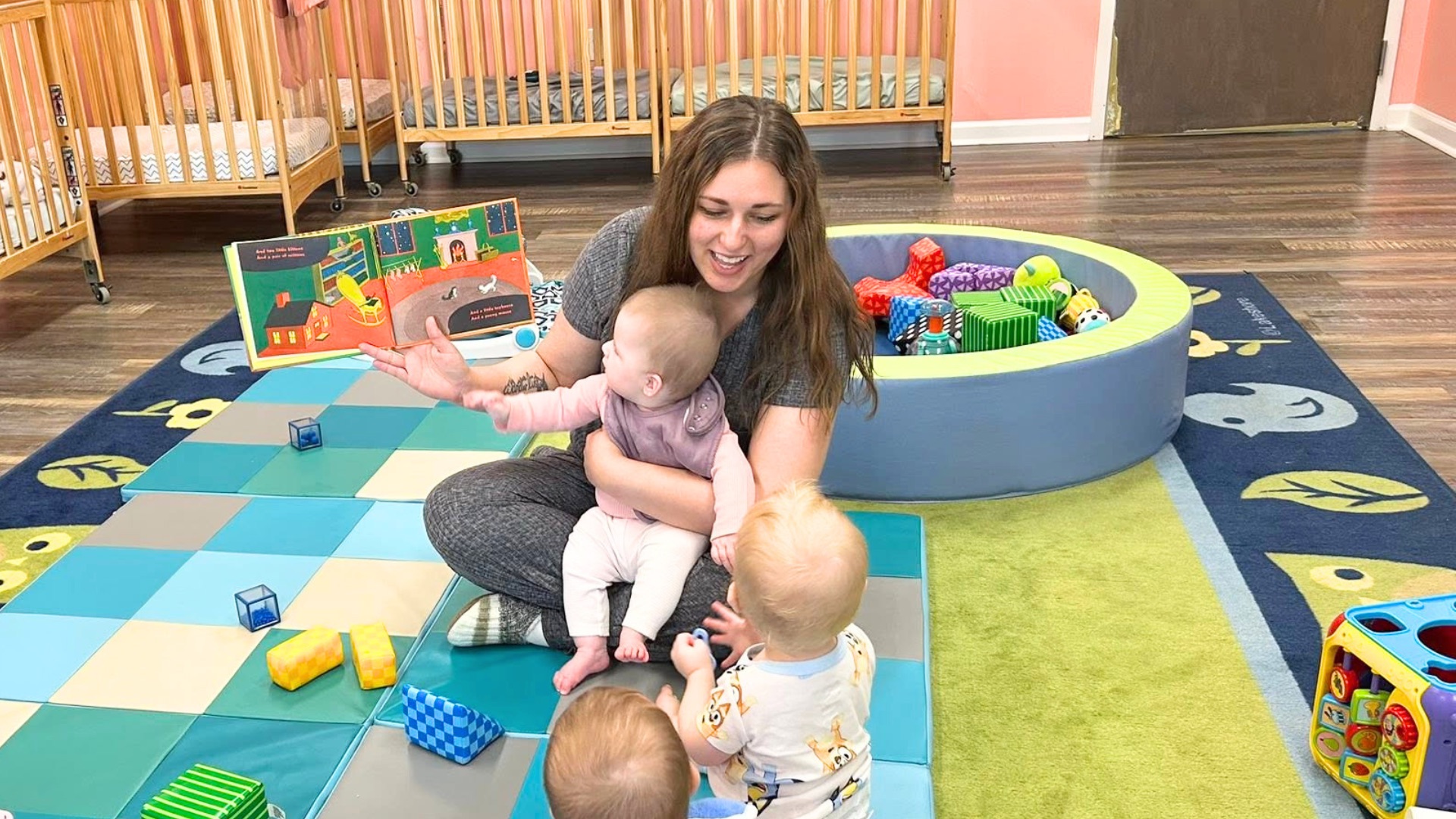 A teacher sitting on a padded play mat reading a picture book to babies in a pink infant room with wooden cribs and a soft ball pit