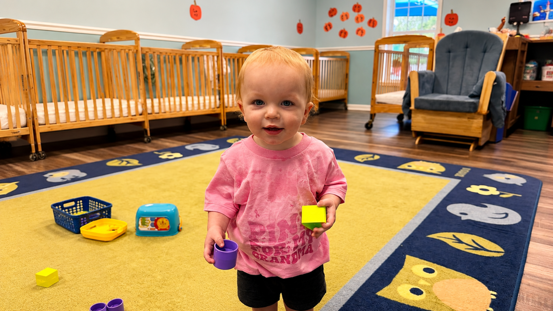 A toddler in a pink shirt standing on a yellow and navy play rug, holding a purple cup and a green block beside a row of wooden cribs