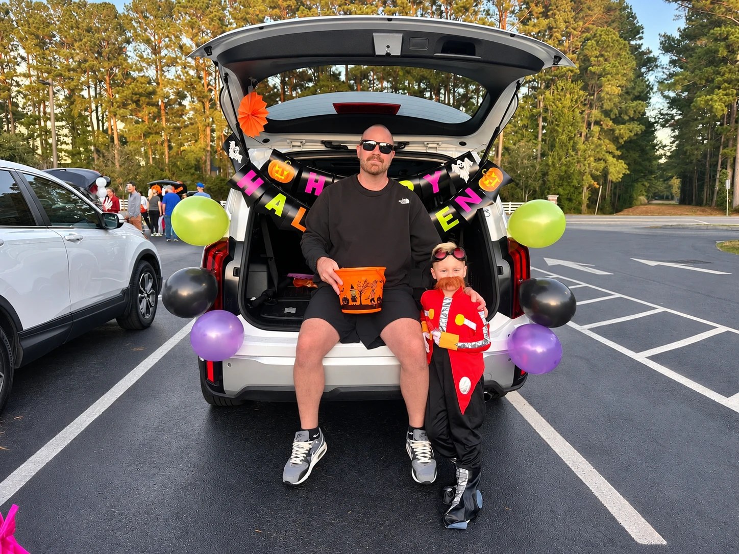A parent and a young child in costume sitting in the back of an SUV with its trunk decorated with Halloween banners and balloons for trunk-or-treating