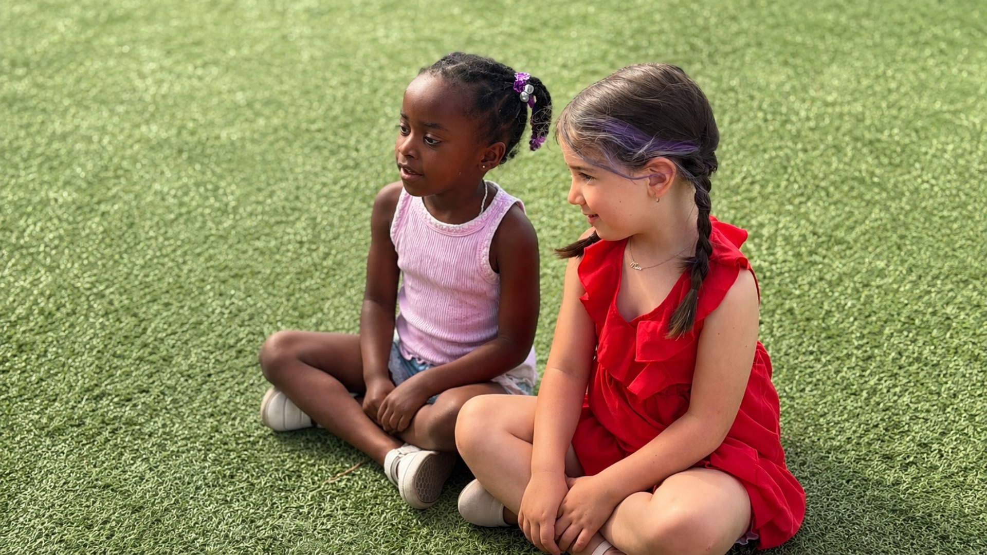 Two school-age girls sitting cross-legged on green turf chatting with each other