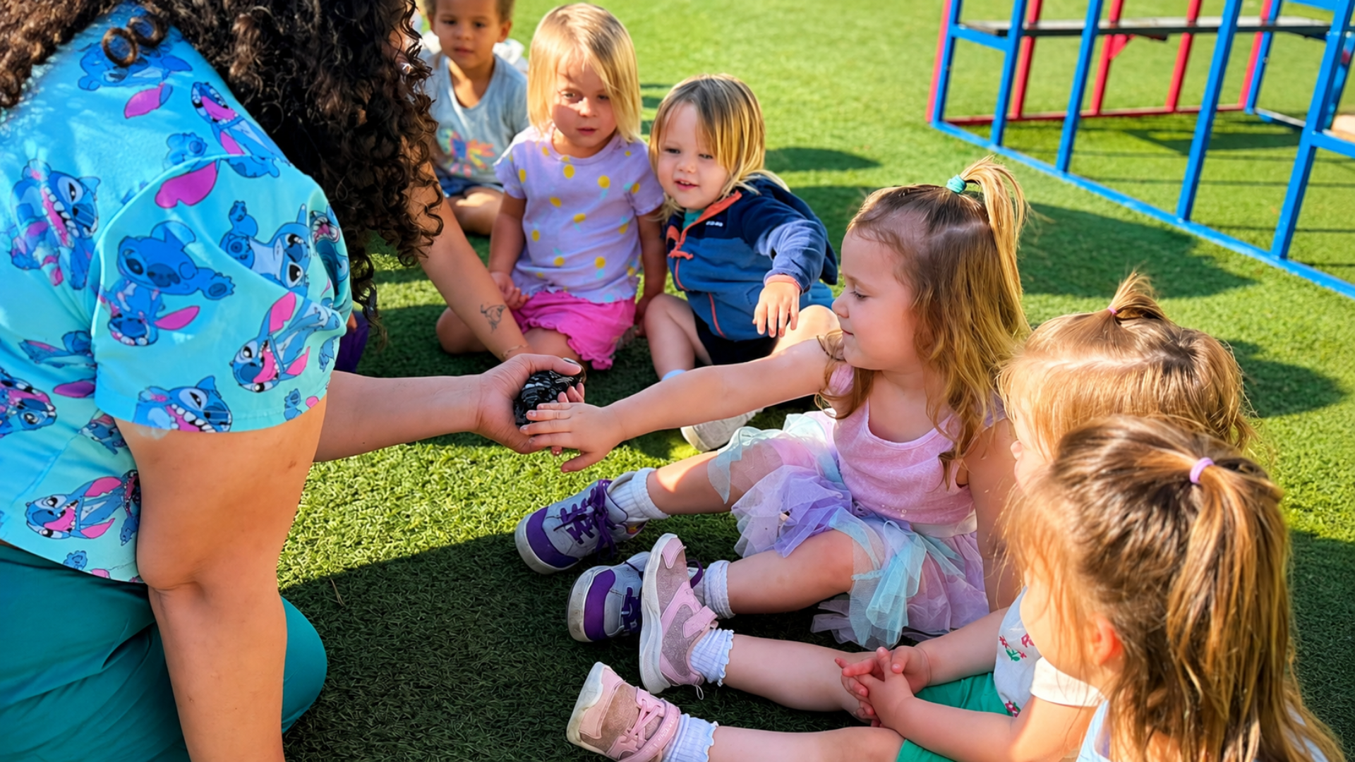 A teacher crouching on the turf playground holding a chick out to a circle of seated preschoolers reaching to touch it