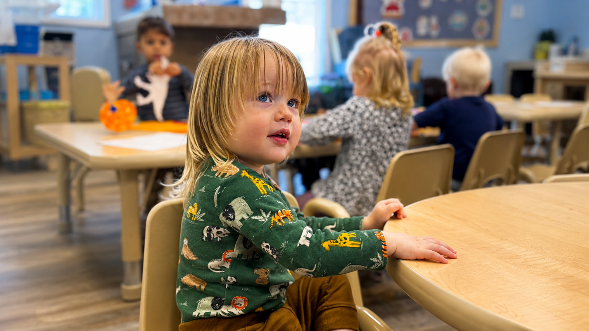 A blond toddler in a green animal-print shirt seated at a wooden classroom table, looking up while classmates work at tables behind