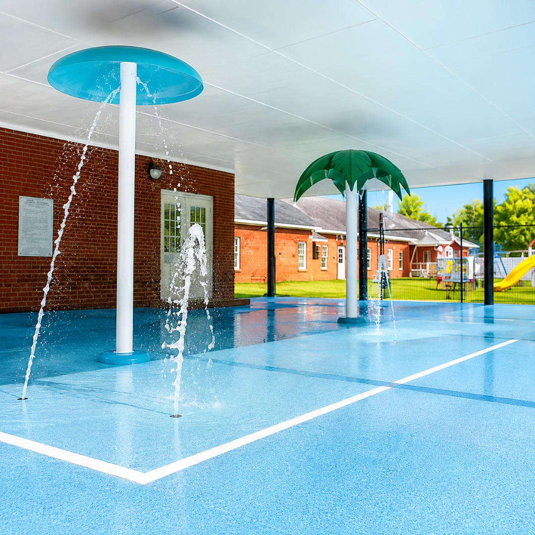 Covered outdoor splash pad with blue rubber flooring, a teal mushroom shower and a green palm-tree sprinkler beside the brick school building and playground