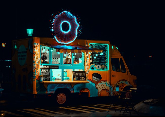 A brightly lit food truck at night with a glowing neon donut sign on top