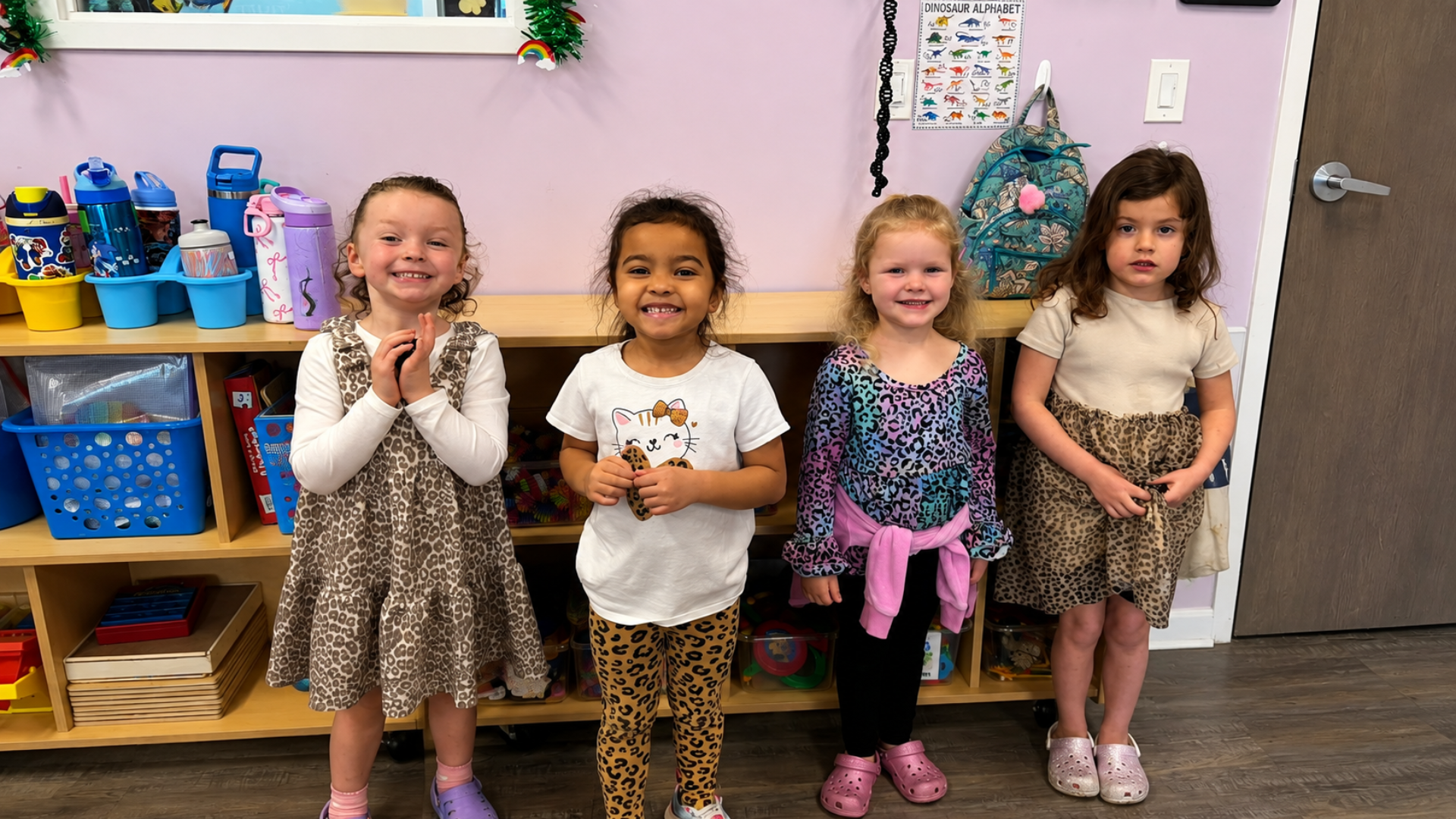 Four preschool girls standing side by side and smiling in front of classroom cubbies stocked with water bottles and bins