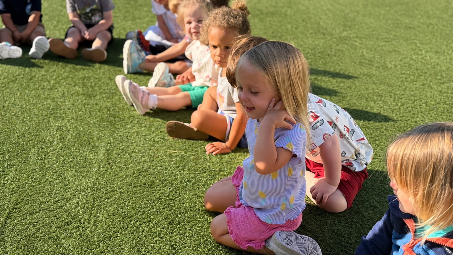 Preschoolers sitting in a row on bright green turf outside in the afternoon sun