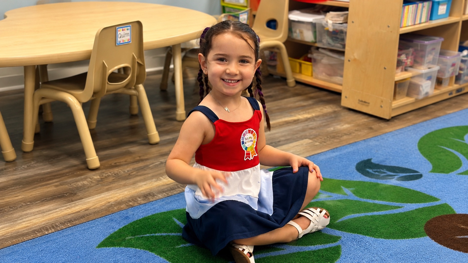 A pre-K girl with braids in a red, white, and navy dress smiling on a leaf-patterned classroom rug beside a kidney table