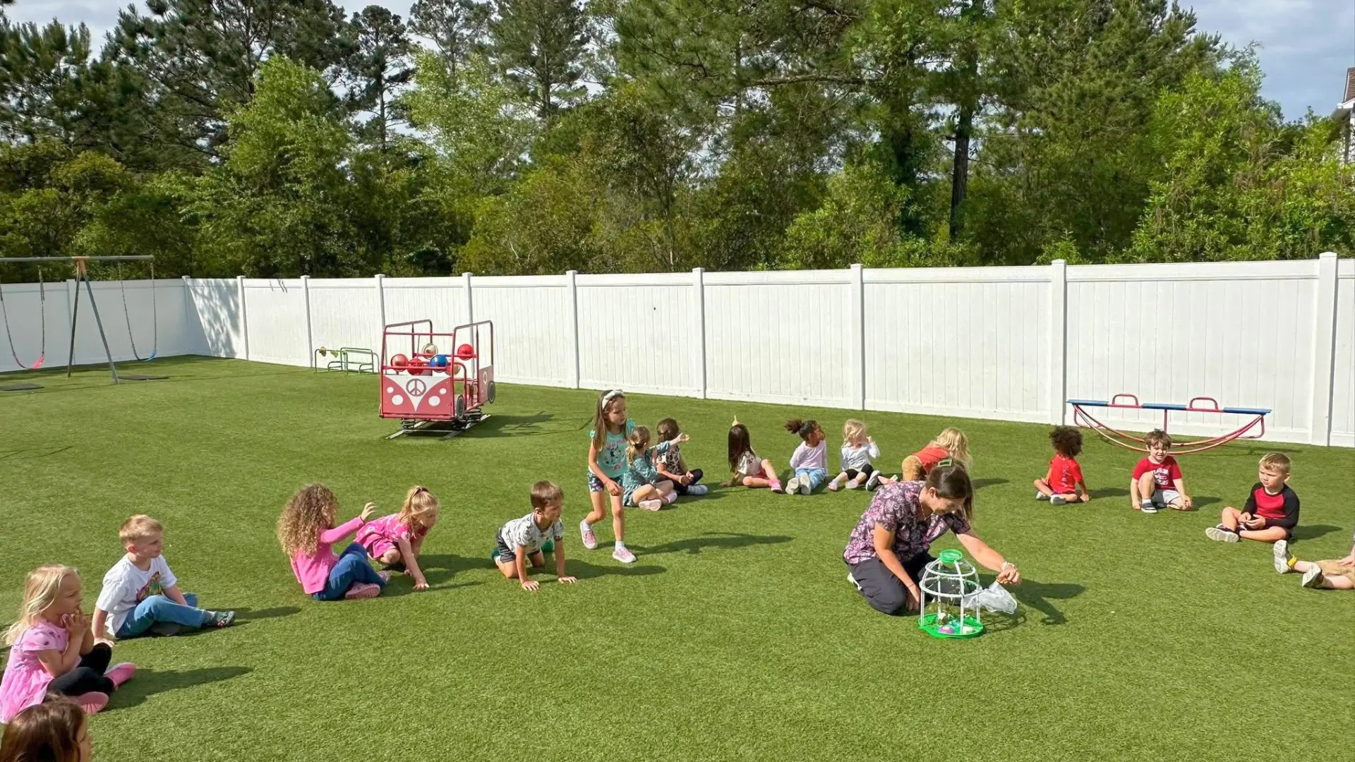 School-age children sitting on the turf playground while a teacher shows them a bug-catching habitat