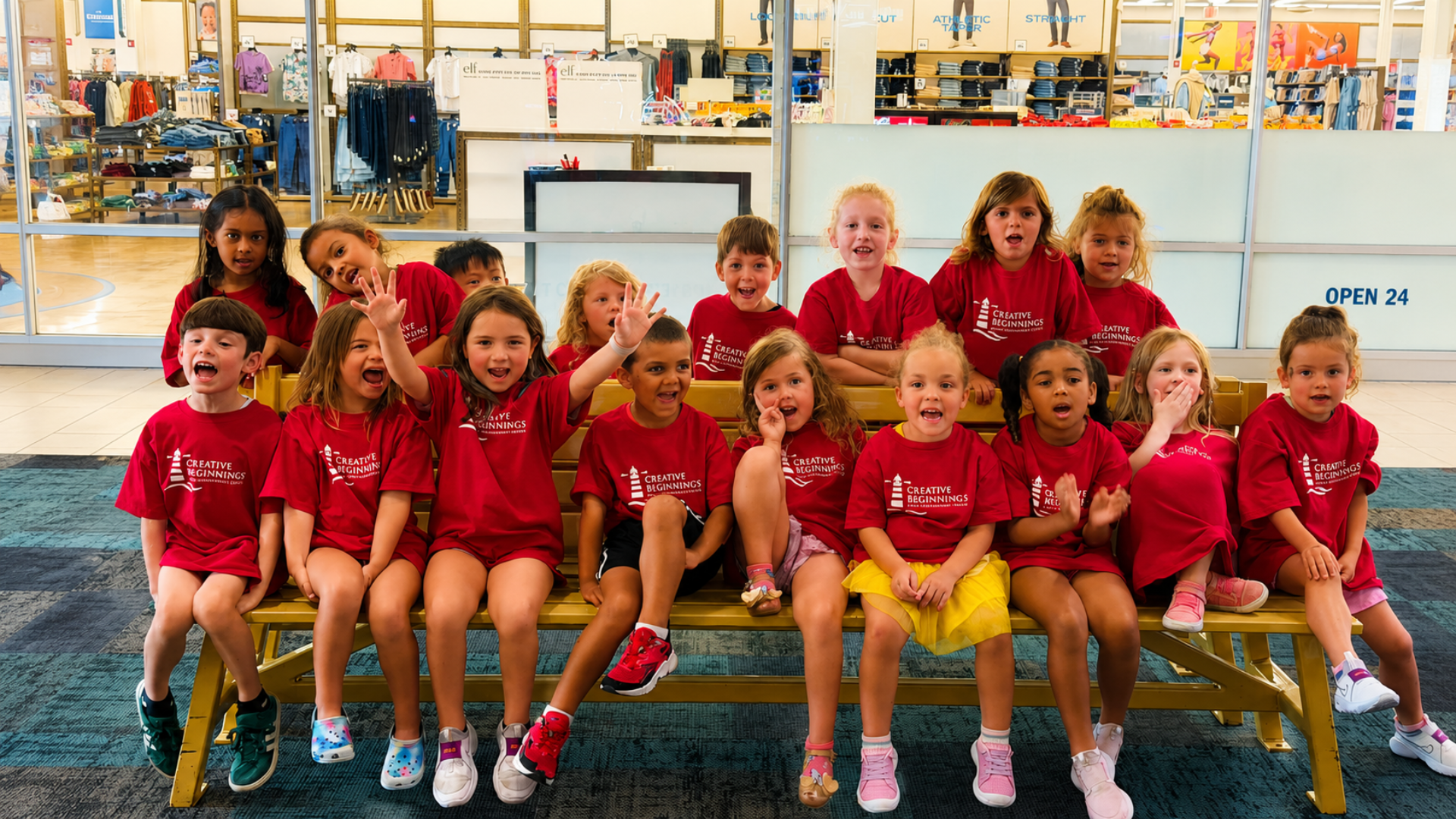 A large group of summer campers in matching red Creative Beginnings T-shirts posing and waving from a bench during a field trip