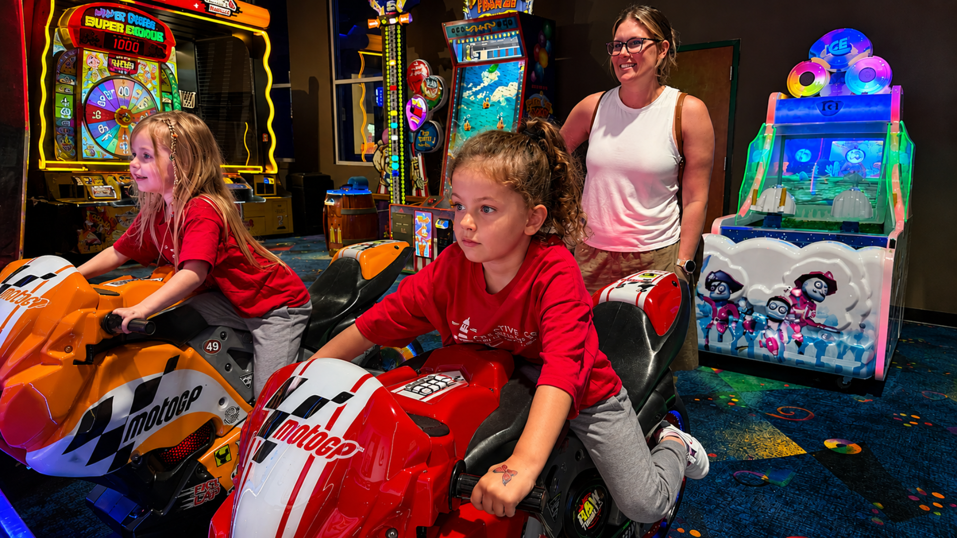 Two campers in red Creative Beginnings shirts riding motorcycle arcade games while a counselor watches from behind