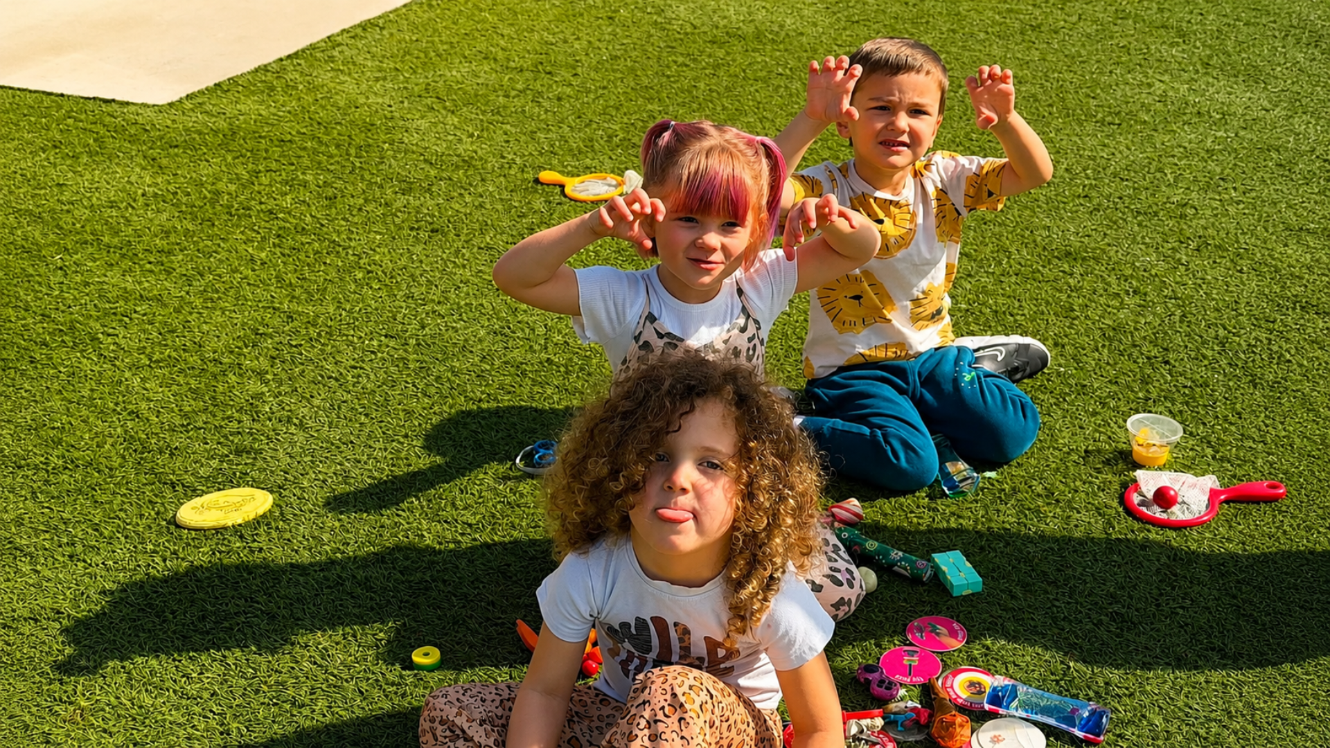 Three pre-K children sitting on the sunny turf playground making playful animal claws at the camera, toys scattered around them
